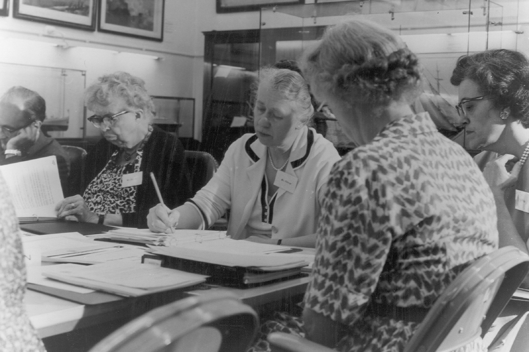 Eleanor Roosevelt and a group of women writing at a shared desk strewn with papers